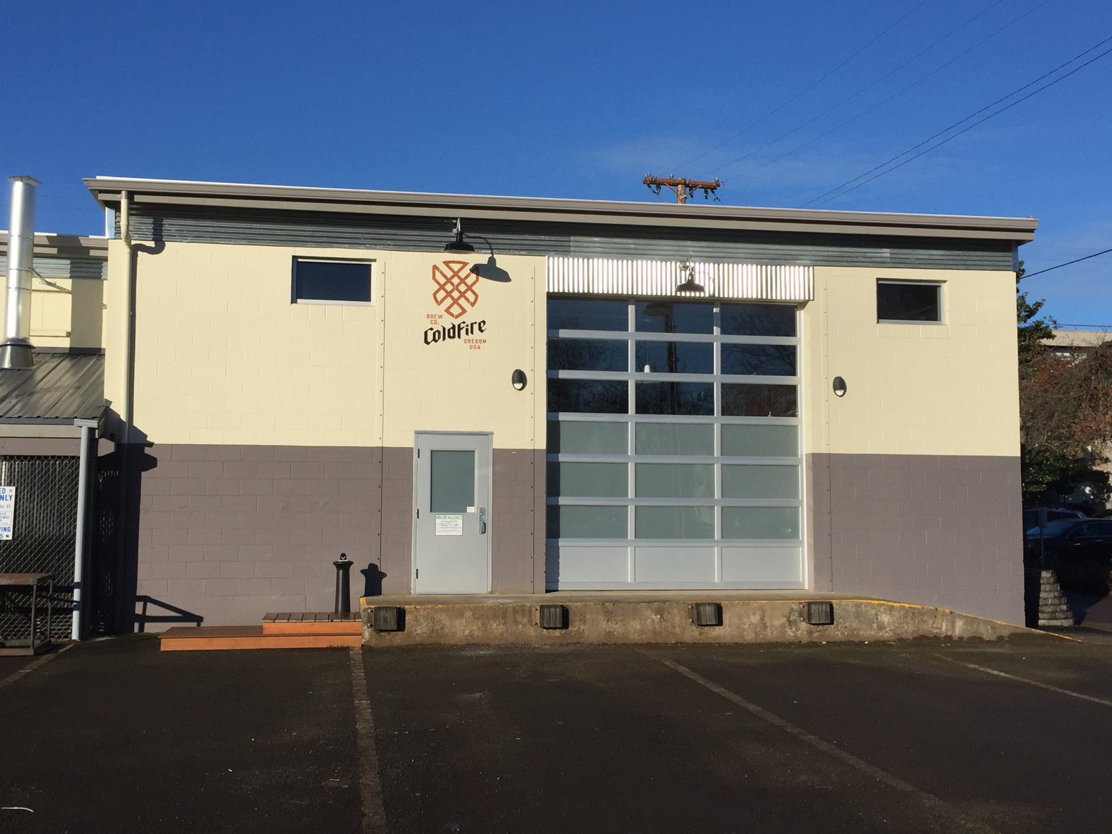 Cream and gray brewery building with a glass roll-up door and brewery logo under a blue sky